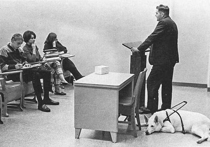 Dr. Eugene Bradfield gives a lecture while Suzie, his seeing-eye dog, rests. (1967 Ah-Mic yearbook)